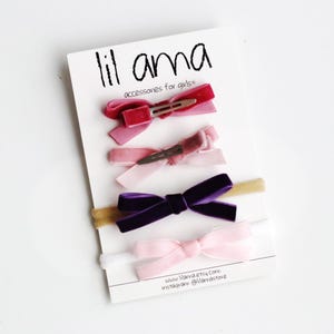 May include: A set of five velvet hair bows in pink, purple, and red, with two matching headbands. The bows are attached to alligator clips. The headbands are made of a thin, stretchy material. The card reads "lil ama accessories for girls" and includes the website and Instagram handle.