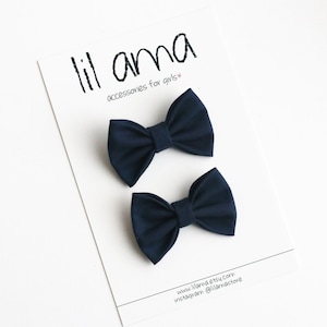 May include: Two navy blue fabric bow hair clips. The bows are tied in a classic bow shape and are attached to a clip. The text "lil ama accessories for girls" is printed on a white card behind the bows.