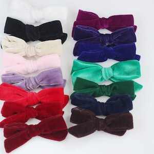 May include: A collection of velvet hair bows in various colors, including white, black, cream, pink, purple, red, burgundy, purple, blue, green, and brown. The bows are arranged in rows on a white background.