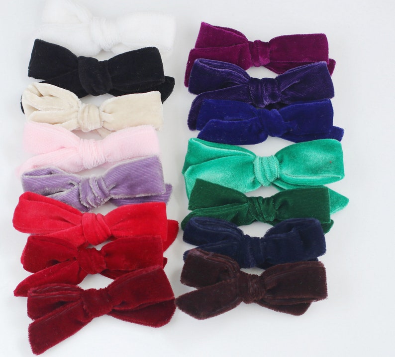 May include: A collection of velvet hair bows in various colors, including white, black, cream, pink, purple, red, burgundy, purple, blue, green, and brown. The bows are arranged in rows on a white background.