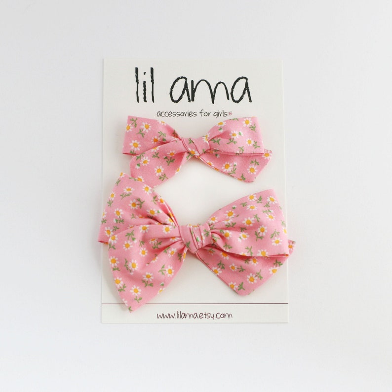 K&ouml;nnte beinhalten: Zwei rosa Haarb&auml;nder mit einem wei&szlig;en G&auml;nsebl&uuml;mchen-Druck. Die Schleifen befinden sich auf einer wei&szlig;en Karte mit dem Text "lil ama accessories for girls*" und "www.lillama.etsy.com".