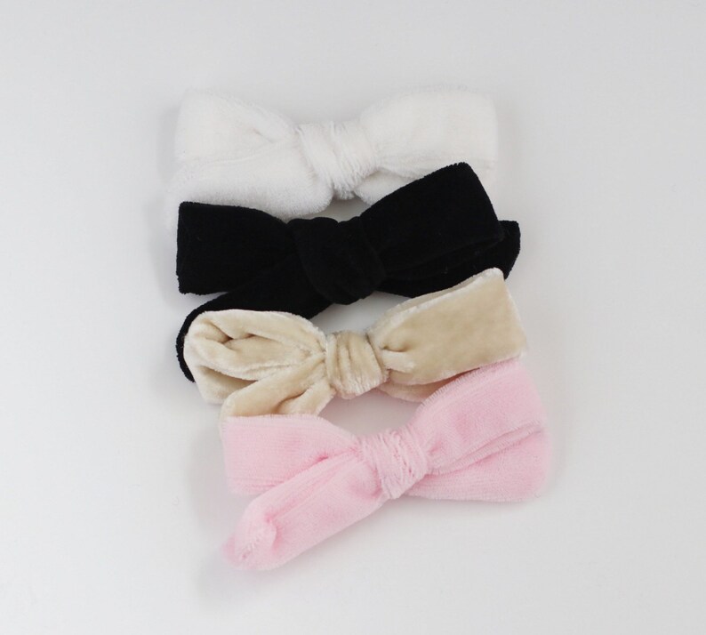 May include: Four velvet bow hair accessories in white, black, beige, and pink. The bows are arranged in a vertical stack, showcasing their soft texture and color variations. These hair bows are a stylish accessory for various hairstyles.