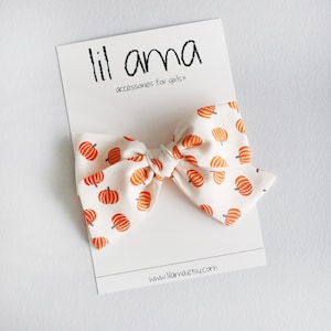 May include: A white fabric bow with a repeating pattern of orange pumpkins. The bow is tied in the center and is attached to a white card with the text "lil ama accessories for girls". The website address www.lilamaetsy.com is also visible.