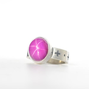 Peut inclure: Bague en argent ornée d'une grosse pierre précieuse ovale magenta avec un motif en étoile à six branches. Le bracelet en argent est orné de découpes en forme de croix. Sur fond blanc.