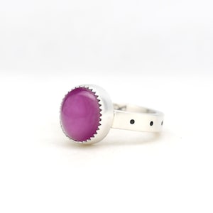 May include: A silver ring featuring a round, cabochon-cut magenta stone set in a serrated bezel. The band is made of smooth silver and is accented with a series of small, evenly spaced black circular inlays along the outer surface of the shank.