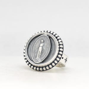 May include: A silver ring featuring a circular face with a beaded border. The center displays an oval medallion of the Miraculous Medal with the text O MARY CONCEIVED WITHOUT SIN PRAY FOR US WHO HAVE RECOURSE TO THEE 1830. The band has a small cross.