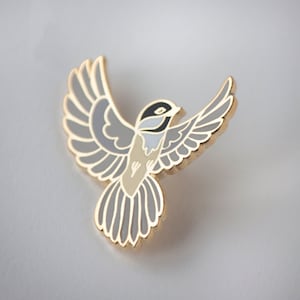 May include: A gold-coloured metal bird pin with outstretched wings. The bird features a design with grey, black, and beige enamel. The pin is a decorative accessory, suitable for jackets, bags, or hats.