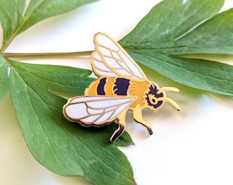 May include: Gold-colored enamel pin of a bee with white wings, a yellow and black striped abdomen, and purple accents. The bee is perched on a green leaf.