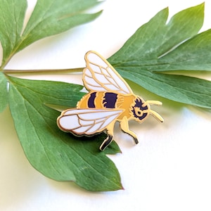 May include: Gold-coloured enamel pin of a bee with white wings, a yellow and black striped abdomen, and purple accents. The bee is perched on a green leaf.