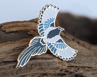Puede incluir: Un pin de esmalte dorado con una urraca azul y blanca en vuelo. El pájaro tiene la cabeza y el cuerpo negros con marcas blancas en las alas.
