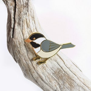 May include: Gold-tone enamel pin of a black-capped chickadee bird with white and grey markings. The bird is perched on a branch with its wings slightly spread.