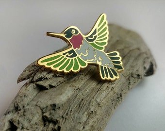 May include: Gold-colored enamel pin of a hummingbird in flight. The hummingbird has green wings, a gray body, a black head, and a red throat.