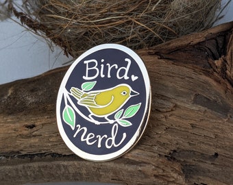 Könnte beinhalten: Eine schwarze und goldene Emaille-Anstecknadel mit einem gelben Vogel, der auf einem grünen Zweig sitzt. Die Anstecknadel trägt die Aufschrift "Bird Nerd" in weißer Kursivschrift.