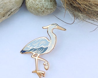 Puede incluir: Un pin de esmalte dorado con un garceta azul de cuello y pico largos. La garceta está de pie sobre una pierna con las alas plegadas. El pin es una representación detallada y realista de una garceta.