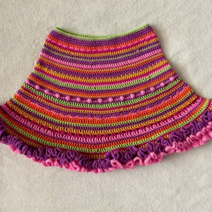 Op de afbeelding: Een kleurrijke gehaakte rok met een gerimpelde zoom. De rok is gemaakt met een verscheidenheid aan felle kleuren, waaronder roze, oranje, geel, groen en paars.