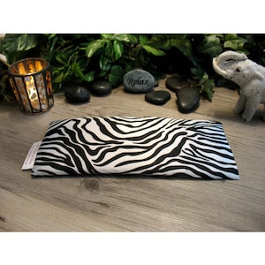 Puede incluir: Una almohada para los ojos rectangular con estampado de cebra en blanco y negro. La almohada está sobre una superficie de madera, con un candelabro, piedras y una pequeña figura de elefante en el fondo. La almohada está diseñada para la relajación.