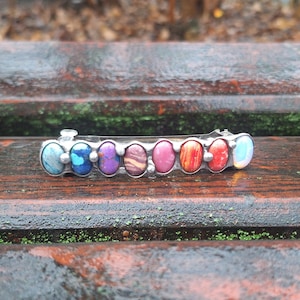 May include: A silver barrette featuring a row of colorful, oval-shaped gemstones. The gemstones include shades of blue, purple, pink, red, and orange. The barrette is resting on a wet, wooden surface.