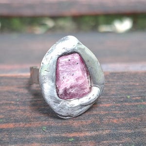 Può includere: Un anello con una pietra preziosa rosa grezza di forma rettangolare, incastonata in una montatura in metallo argentato dalla consistenza irregolare. La superficie metallica presenta segni di spazzolatura visibili e una finitura opaca.