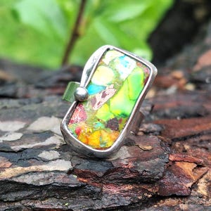 Op de afbeelding: Een zilveren ring met een rechthoekige, veelkleurige steen. De steen heeft tinten groen, geel, blauw en rood. De ring is geplaatst op een getextureerd, bruin oppervlak, met een vage groene achtergrond.