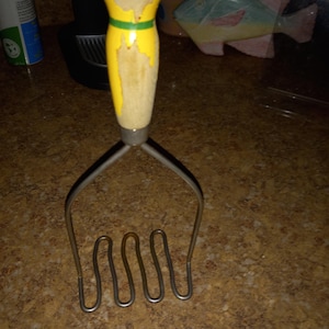 May include: A vintage potato masher with a yellow handle and a green stripe. The handle shows signs of wear, with the yellow paint chipped in places. The masher's metal head has a wavy design.
