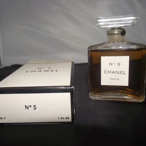 May include: A rectangular glass perfume bottle filled with amber liquid, featuring a clear stopper and a white label reading No 5 CHANEL PARIS. Beside it sits a white cardboard box with black trim, labeled No 5 CHANEL and 1 FL OZ.