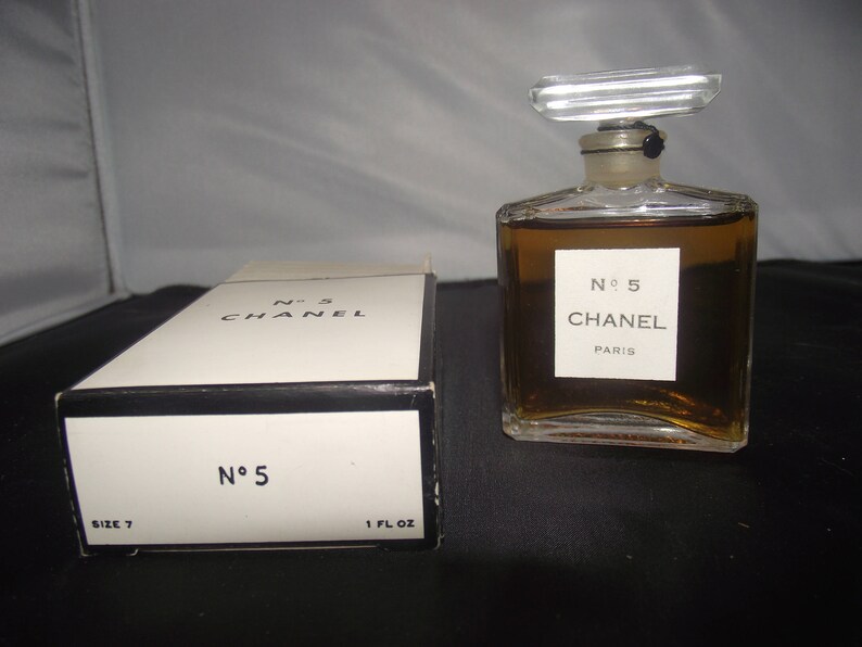 May include: A rectangular glass perfume bottle filled with amber liquid, featuring a clear stopper and a white label reading No 5 CHANEL PARIS. Beside it sits a white cardboard box with black trim, labeled No 5 CHANEL and 1 FL OZ.
