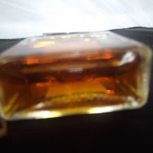 May include: A rectangular glass bottle filled with an amber-colored liquid. The bottle has clear, thick glass walls and a flat base. The liquid inside is translucent and dark orange in tone, filling the container almost to the top edge.