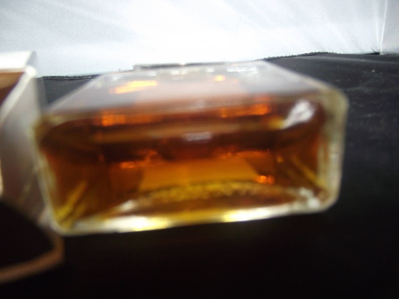 May include: A rectangular glass bottle filled with an amber-colored liquid. The bottle has clear, thick glass walls and a flat base. The liquid inside is translucent and dark orange in tone, filling the container almost to the top edge.