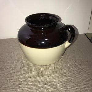 May include: A ceramic crock with a rounded body and a single side handle. The upper portion of the vessel features a dark brown glossy glaze, while the lower portion is finished in a smooth, light cream color. The rim is wide and circular.
