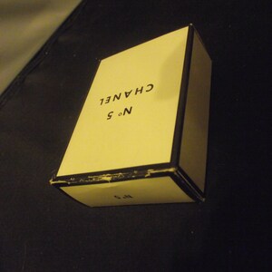 May include: A rectangular cream-colored box with black trim and edges. The top surface features the black text CHANEL and N&deg; 5. The front side of the box also displays the text N&deg; 5 in black. The box appears to be a container for a fragrance product.