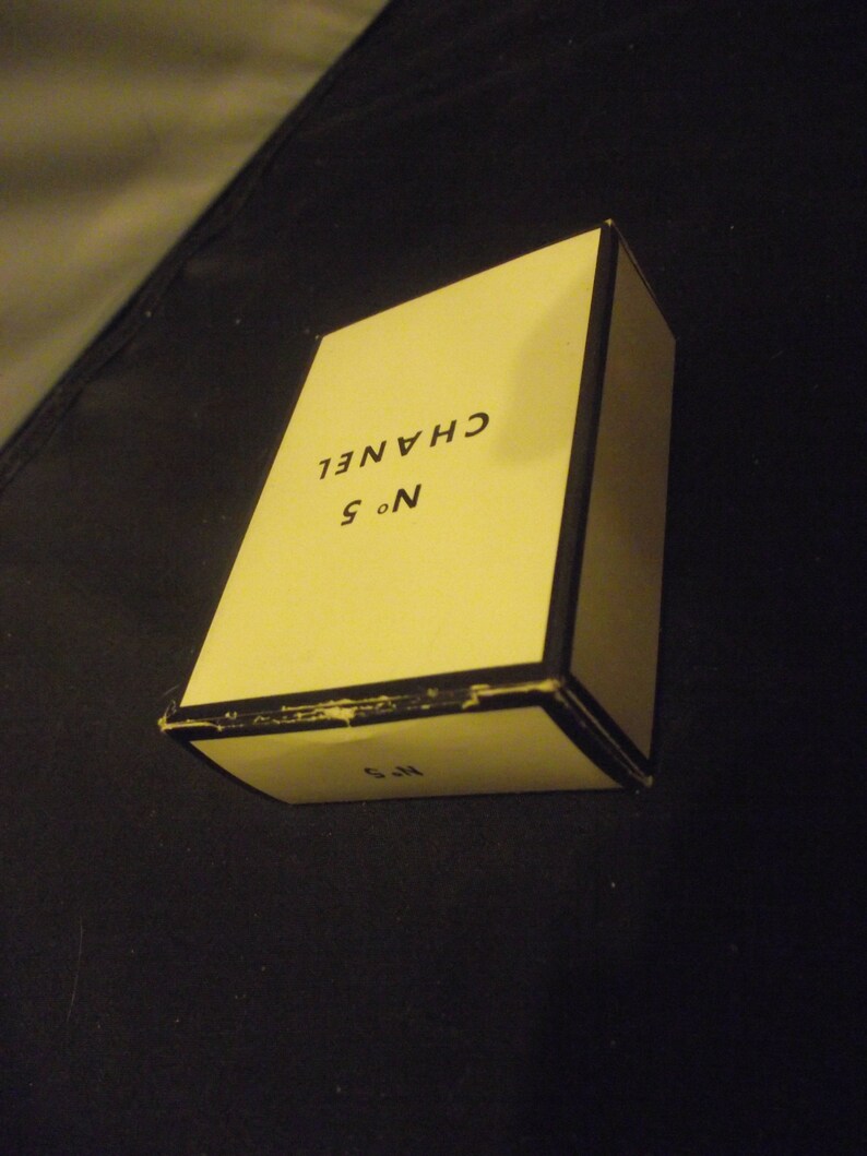 May include: A rectangular cream-colored box with black trim and edges. The top surface features the black text CHANEL and N&deg; 5. The front side of the box also displays the text N&deg; 5 in black. The box appears to be a container for a fragrance product.
