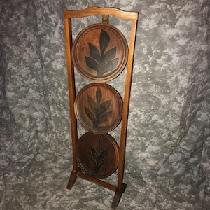 May include: A tall wooden stand featuring three vertically stacked circular wooden plaques. Each plaque is carved with a dark brown leaf design against a lighter wood background. The frame is constructed from light brown wood with a simple base.