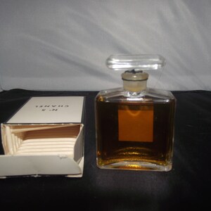May include: A rectangular glass perfume bottle filled with amber liquid, featuring a clear stopper and a blank square label. Beside it sits an open white cardboard box with the text N&deg;5 CHANEL printed in black on the top flap of the packaging.