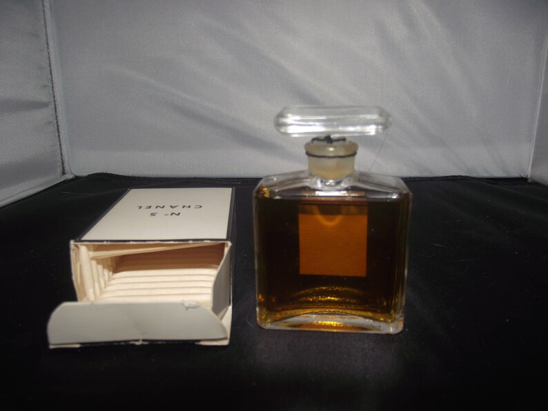 May include: A rectangular glass perfume bottle filled with amber liquid, featuring a clear stopper and a blank square label. Beside it sits an open white cardboard box with the text N&deg;5 CHANEL printed in black on the top flap of the packaging.