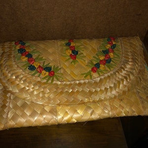 Pu&ograve; includere: Una pochette in paglia intrecciata con chiusura a patta. La patta &egrave; decorata con un motivo floreale rosso, giallo, blu e verde. La pochette &egrave; rettangolare e sembra fatta a mano.