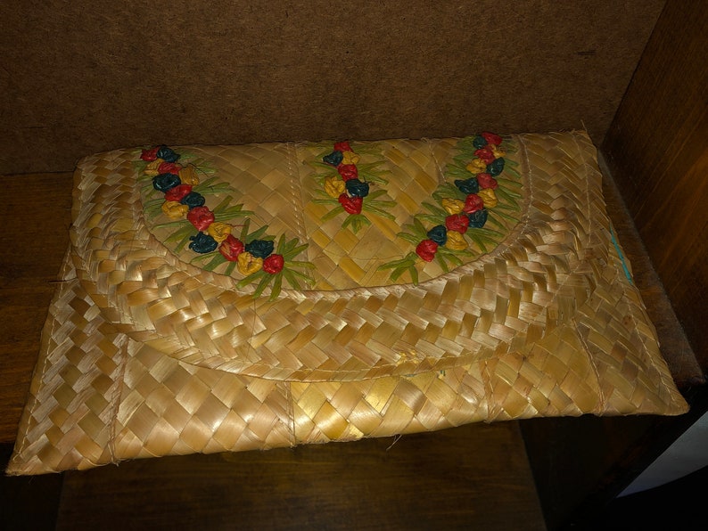 Pu&ograve; includere: Una pochette in paglia intrecciata con chiusura a patta. La patta &egrave; decorata con un motivo floreale rosso, giallo, blu e verde. La pochette &egrave; rettangolare e sembra fatta a mano.