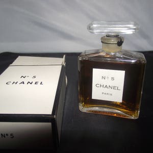 May include: A clear glass perfume bottle filled with amber liquid, featuring a rectangular label that reads No 5 CHANEL PARIS and a clear rectangular stopper. Beside it sits a white rectangular box with black trim labeled No 5 CHANEL.