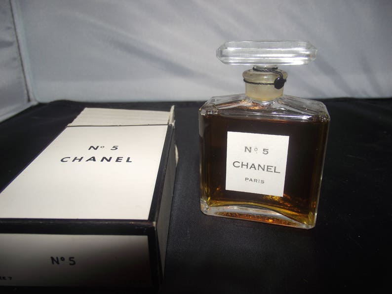 May include: A clear glass perfume bottle filled with amber liquid, featuring a rectangular label that reads No 5 CHANEL PARIS and a clear rectangular stopper. Beside it sits a white rectangular box with black trim labeled No 5 CHANEL.