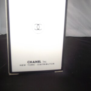 May include: A white rectangular box with a black border featuring the black Chanel interlocking C logo centered near the top. The text CHANEL Inc. NEW YORK - DISTRIBUTOR is printed in black at the bottom of the front face of the packaging.