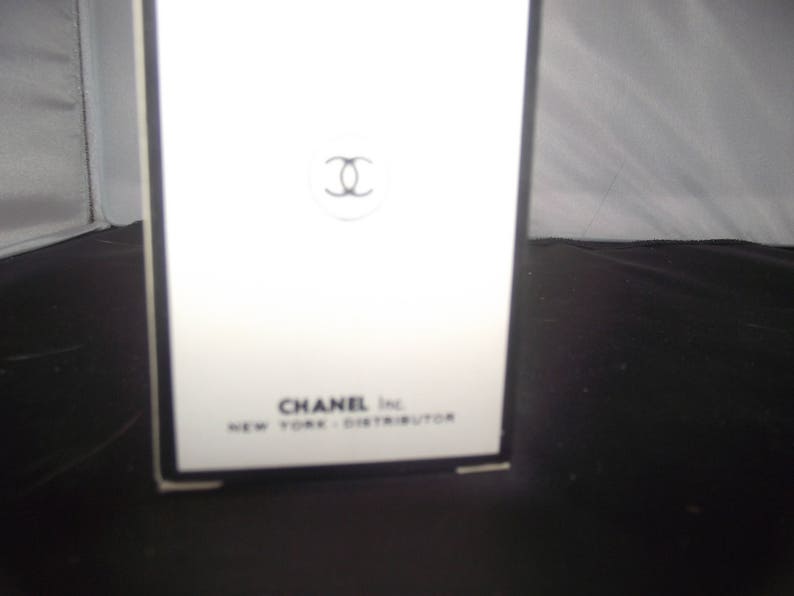 May include: A white rectangular box with a black border featuring the black Chanel interlocking C logo centered near the top. The text CHANEL Inc. NEW YORK - DISTRIBUTOR is printed in black at the bottom of the front face of the packaging.