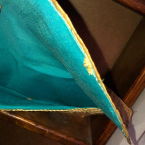 Pu&ograve; includere: Primo piano di una scatola di legno con fodera in tessuto turchese. Il tessuto ha un bordo dorato ed &egrave; parzialmente tirato indietro, rivelando l'interno. Il legno ha una finitura scura e ricca.