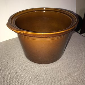 May include: A brown ceramic pot with a rounded shape and a wide opening. The pot has a glossy finish and a darker brown interior. It features two small handles on the sides and sits on a neutral-colored surface.