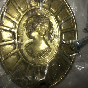 May include: Oval-shaped brass cabinet pull with a classical female profile in the center, surrounded by a sunburst design. The words "Pat. Pend." and "USA" are visible. Two silver screws are included.