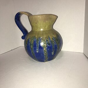 May include: A ceramic pitcher with a rounded body and a curved handle. The upper portion features a muted tan and olive green glaze, while the lower section displays a vibrant blue glaze that drips downward over the green base in vertical streaks.