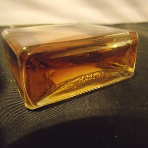 May include: A rectangular glass perfume bottle filled with amber-colored liquid. The base of the clear glass bottle features embossed text that reads "BOTTLE MADE IN FRANCE". The bottle has sharp, defined corners and a smooth, transparent surface.