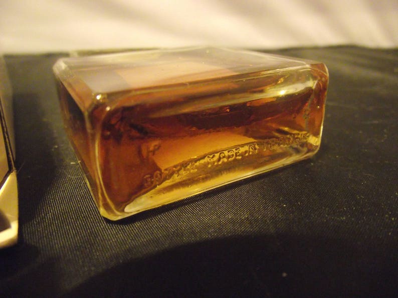 May include: A rectangular glass perfume bottle filled with amber-colored liquid. The base of the clear glass bottle features embossed text that reads "BOTTLE MADE IN FRANCE". The bottle has sharp, defined corners and a smooth, transparent surface.
