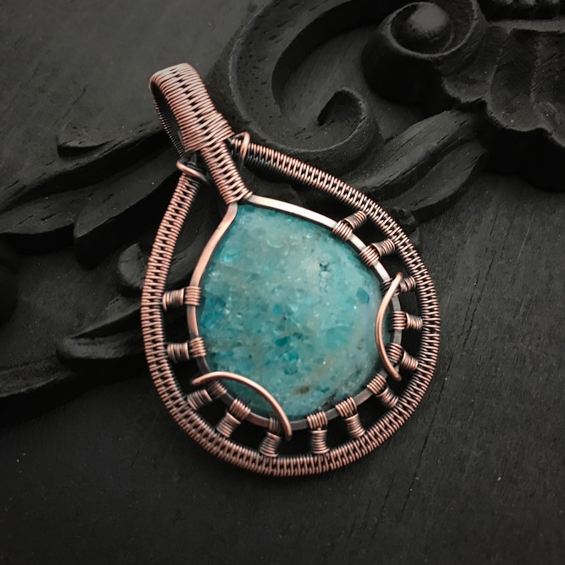 K&ouml;nnte beinhalten: Ein mit Kupferdraht umwickelter Anh&auml;nger mit einem blaugr&uuml;nen Edelstein. Der Anh&auml;nger hat die Form einer Tr&auml;ne und ein filigranes Drahtdesign.