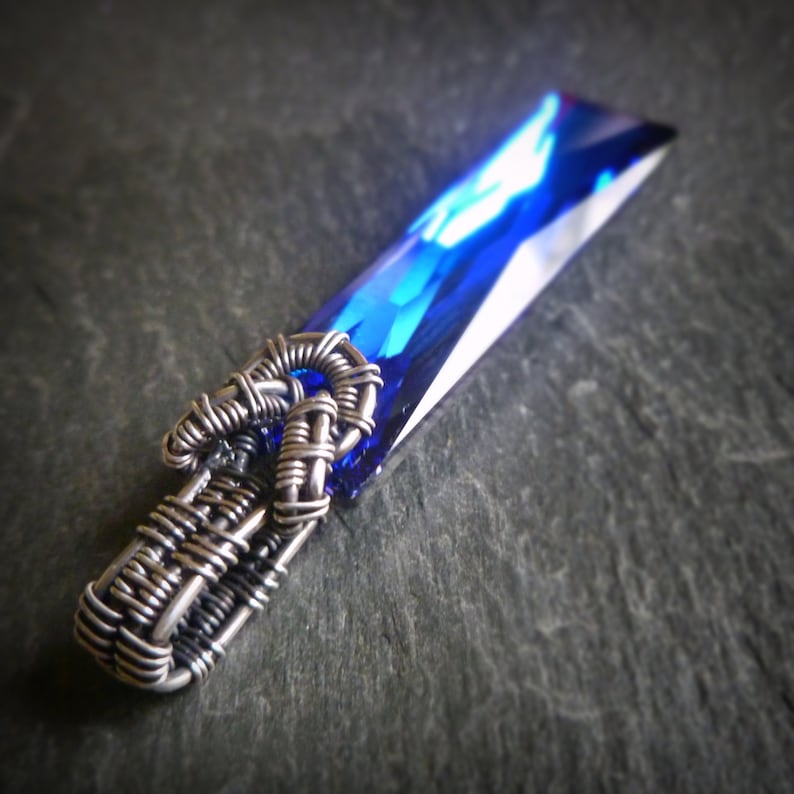 Puede incluir: Un colgante de alambre plateado envuelto alrededor de un cristal azul. El cristal es facetado y tiene una forma rectangular.