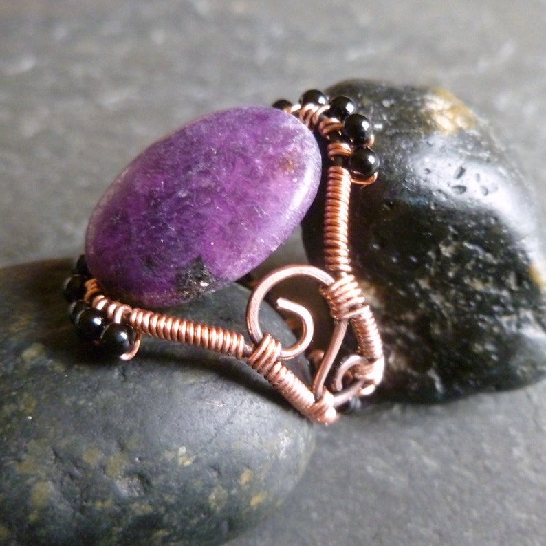 Op de afbeelding: Een ring met een paarse edelsteen, koperdraad en zwarte kralen. De ring is handgemaakt en heeft een uniek ontwerp.