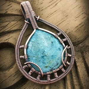 K&ouml;nnte beinhalten: Ein mit Kupferdraht umwickelter Anh&auml;nger mit einem hellblauen Edelstein. Der Anh&auml;nger hat die Form einer Tr&auml;ne und ein filigranes Drahtdesign.